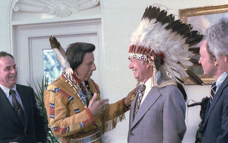A photograph showing Iron Eyes Cody, a known pretendian, in Plains-style regalia. He is standing next to President Jimmy Carter, who is wearing an eagle feather headdress and smiling.