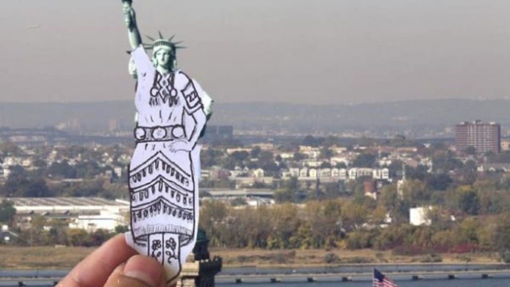 A photo of an ink drawing of a jingle dress held in front of the statue of liberty